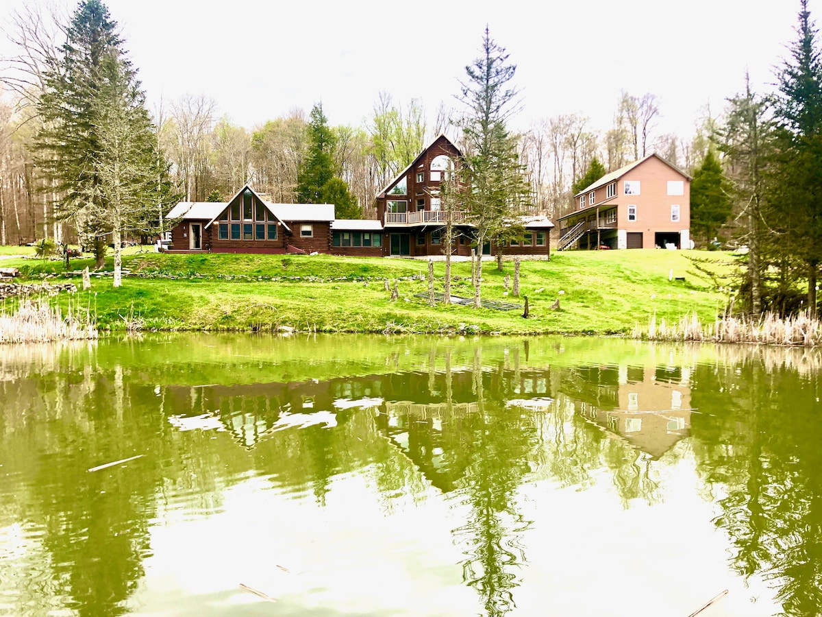 A view of the property reflects in the tranquil pond. The spacious multi-level house features large windows and a blend of wooden and modern architecture, surrounded by well-maintained green lawns and tall trees. Nature creates a serene backdrop to the inviting retreat.