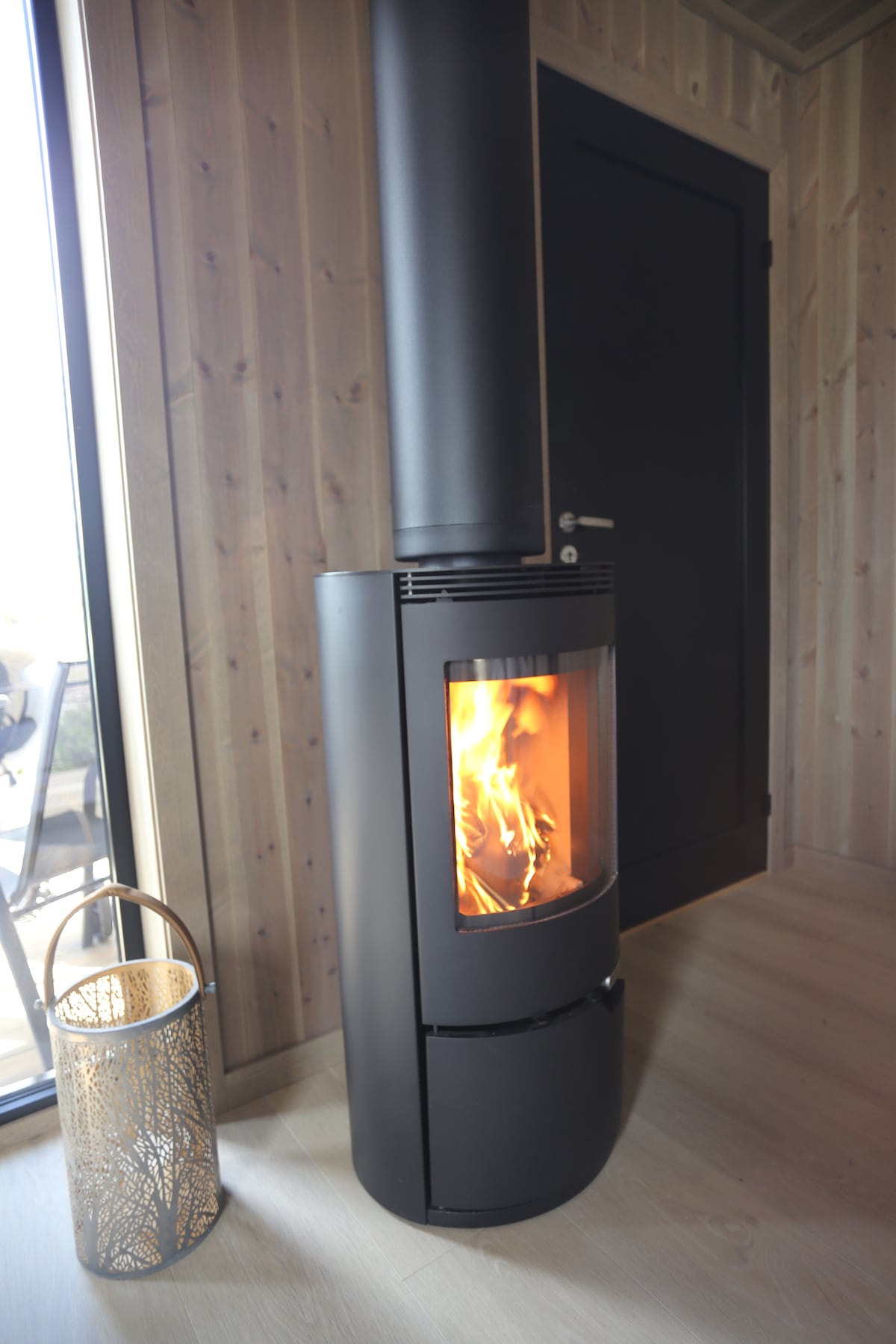 A modern black wood stove is positioned in a corner, with flames flickering inside the glass front. Natural wood paneling surrounds the stove, and a decorative lantern sits nearby, adding warmth to the space.