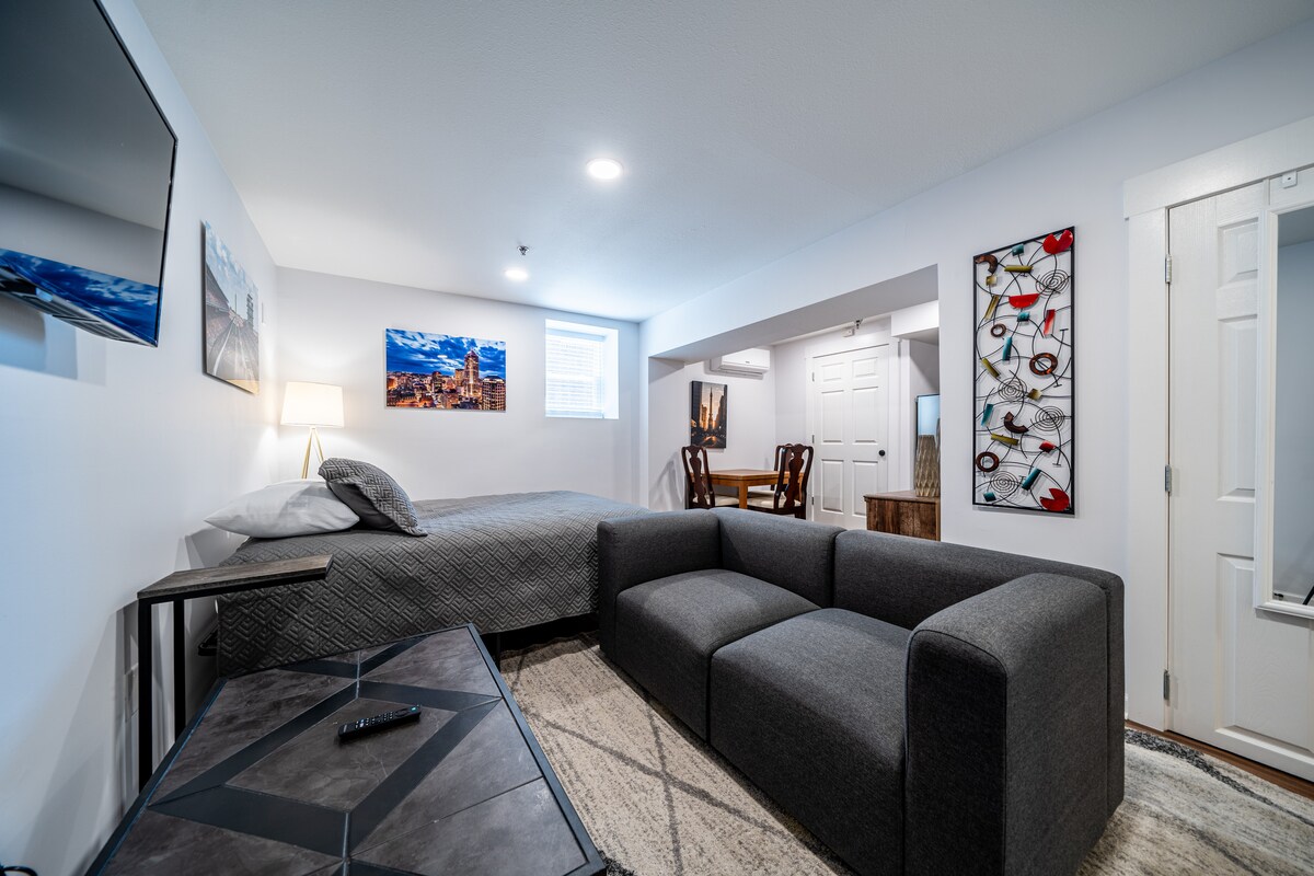 A studio apartment is shown, featuring a spacious bed against a white wall, complemented by artwork. A modern gray sectional sofa is positioned near a coffee table, while a wall-mounted TV is visible. Natural light enters through a window with white blinds.