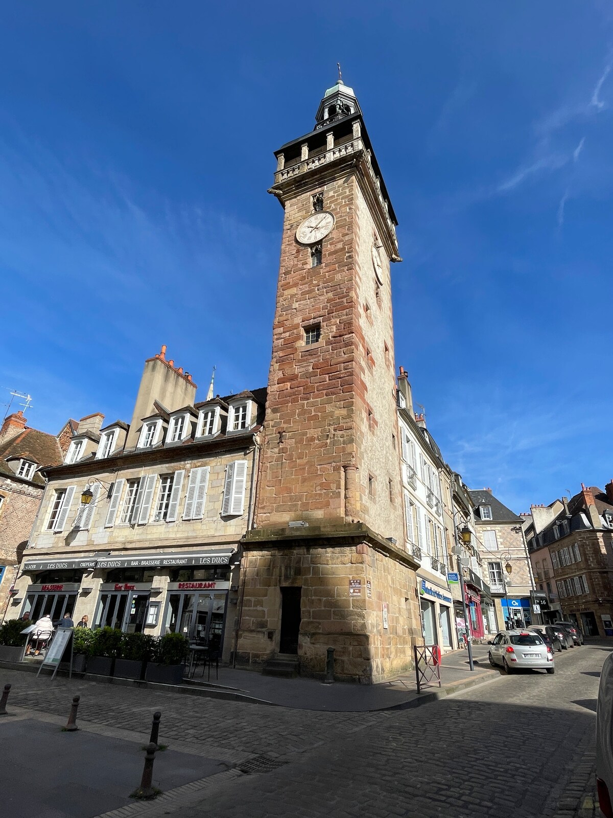 A historic stone clock tower stands tall in a bustling street. Classic architectural features include arched windows and a clock face at the top. Surrounding buildings exhibit traditional design elements, and parked cars line the cobblestone street under a clear blue sky.