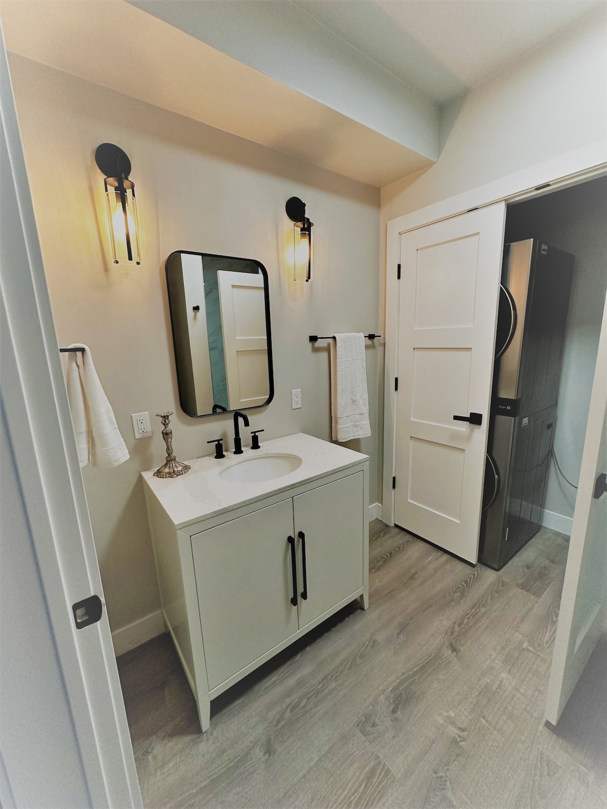 A modern bathroom is presented with a sleek vanity featuring a white countertop and a round mirror above. Soft lighting is provided by sconces on the wall. A door to the right leads to a closet with a washer and dryer, enhancing functionality.