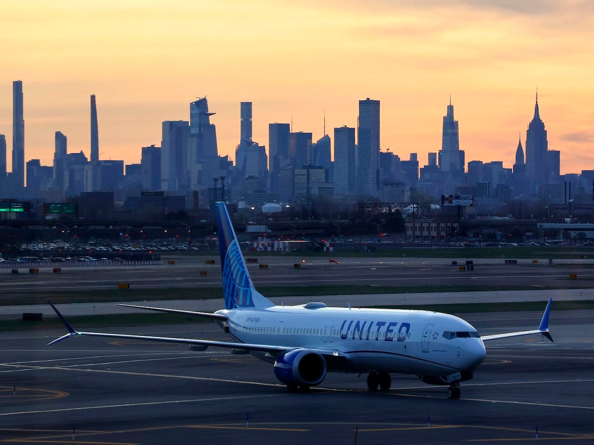 An airplane is positioned on the runway at sunset, with the skyline of New York City in the background. The iconic buildings, including the Empire State Building, are silhouetted against a vibrant sky transitioning from orange to purple.