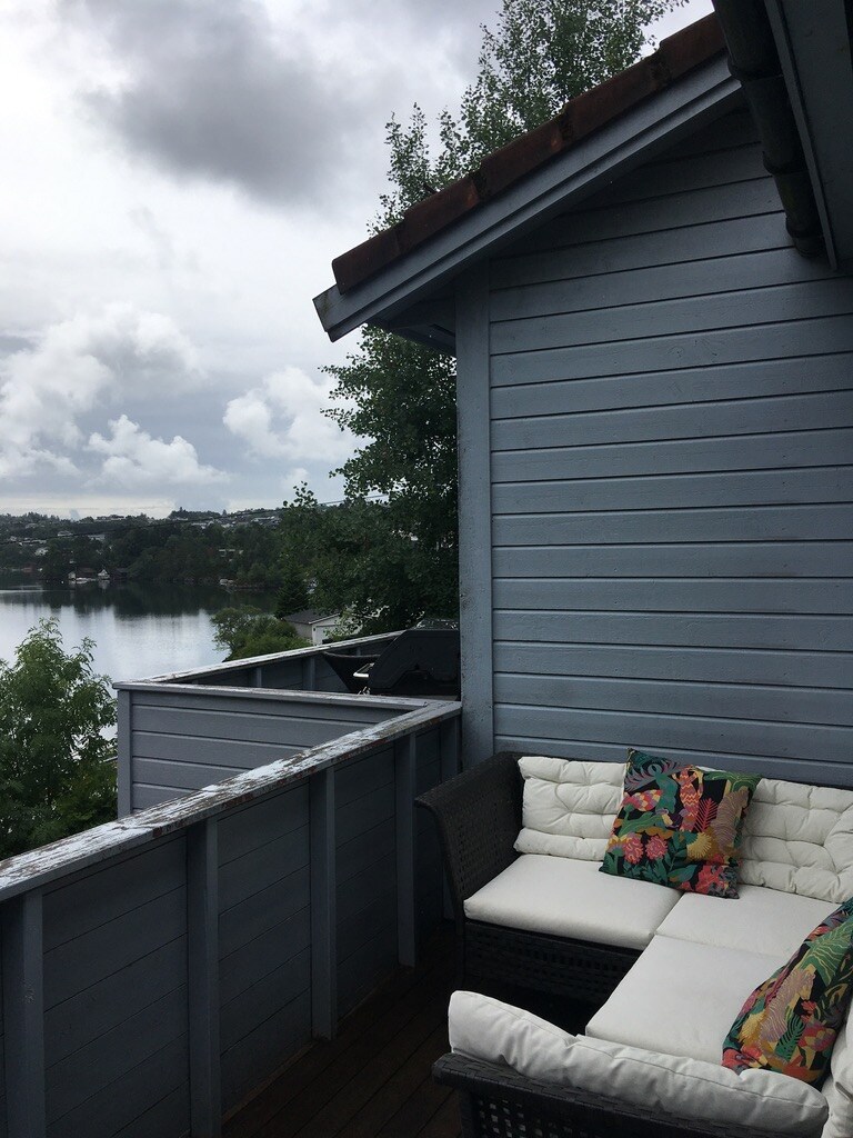 A balcony is visible with a cozy seating area featuring a dark wicker sofa adorned with vibrant, patterned cushions. The serene view of the water and surrounding greenery can be seen in the background, under a cloudy sky.