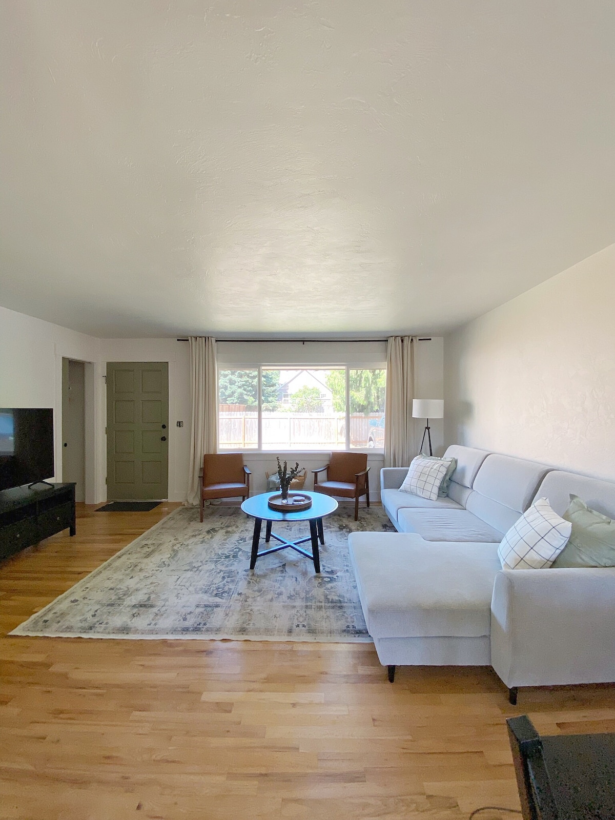 A bright living room features a light-colored sofa with decorative pillows, complemented by two warm-toned armchairs. A round coffee table sits centrally on a soft area rug. Large windows allow natural light to fill the space, with sheer curtains gently framing the view.