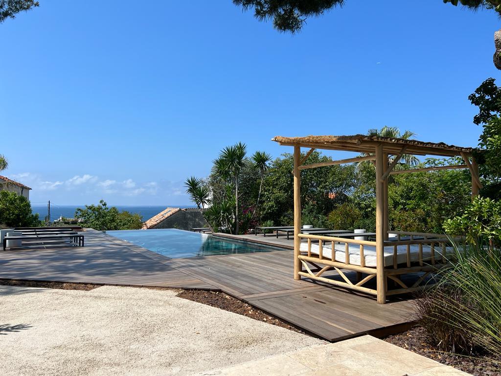 An outdoor space features a wooden deck surrounding a heated swimming pool with a view of the ocean. A shaded lounge area with a canopy is present, accompanied by several seating options, set against a backdrop of lush greenery and palm trees under a clear blue sky.