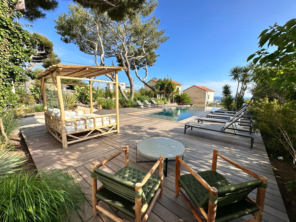 A wooden deck surrounds a serene heated pool, with comfortable lounge chairs positioned for relaxation. A shaded seating area presents a cozy lounging spot. Lush greenery and palm trees add a refreshing touch, enhancing the outdoor ambiance and providing a tranquil escape.