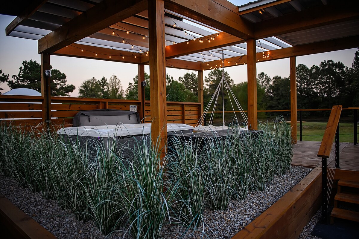 A shaded wooden structure features comfortable seating and a hot tub, surrounded by decorative gravel and lush green grass. Soft string lights are gracefully hung above, adding a serene touch to the outdoor area, which overlooks the natural landscape of the property.