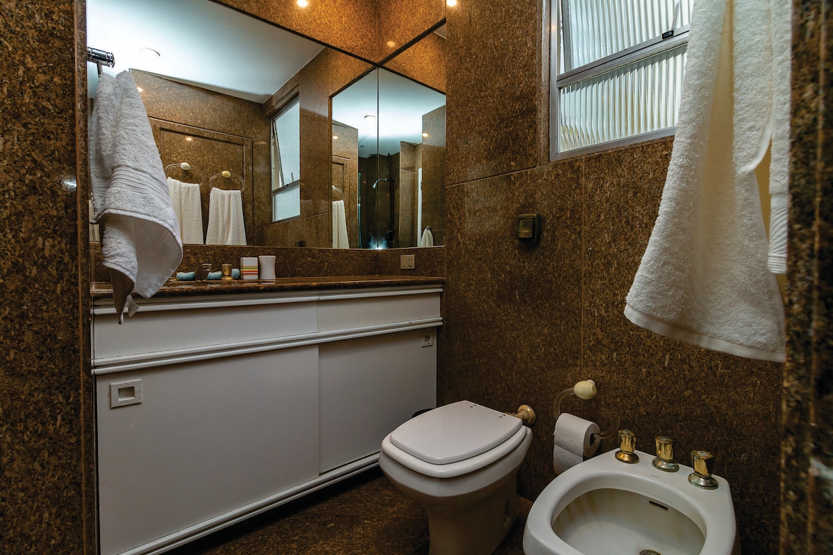 The bathroom is finished with elegant granite surfaces, featuring a large mirror above the sink area. A bidet and toilet are positioned adjacent to each other, with towel bars holding white towels. Natural light enters through a frosted window, enhancing the space.