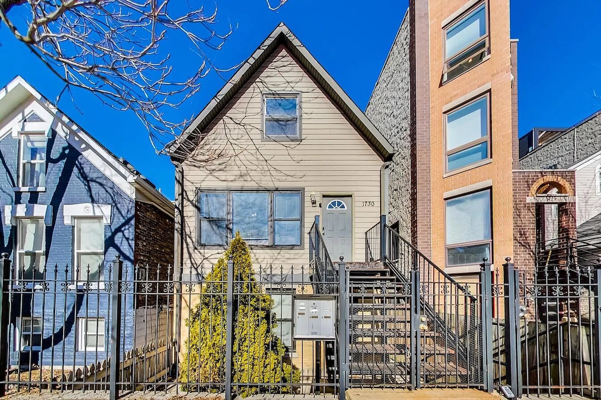 A charming two-story building with a light brown exterior is framed by a black wrought-iron fence. A well-maintained staircase leads to the front entrance, while the neighborhood features a mix of architectural styles. Lush greenery is visible alongside the pathway.