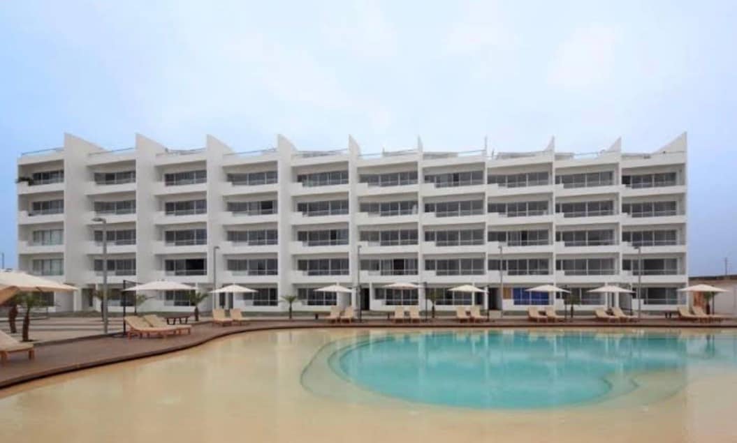 A modern multi-story building is depicted, featuring multiple balconies and large windows. The photo showcases a curved pool area surrounded by lounge chairs and umbrellas, offering a space for relaxation. The overall layout emphasizes an inviting atmosphere for family gatherings.