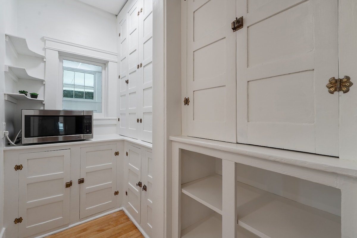 A bright and airy kitchen space is presented, featuring white cabinetry with decorative brass hardware. A compact microwave is visible on the countertop, while the window allows natural light to illuminate the area. Open shelving and spacious cabinets offer ample storage options.