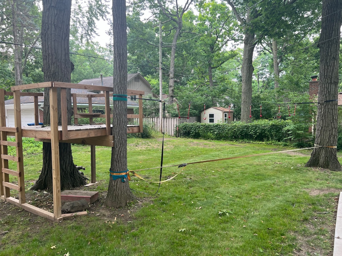 A backyard scene showcases a wooden play structure positioned between two trees. A slackline is visible extending between the trees in the grassy area. Lush greenery provides a natural backdrop, while a small shed is partially seen in the distance.