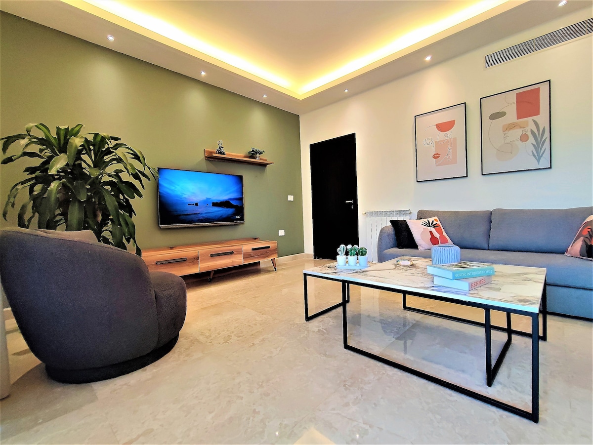 A spacious living room is highlighted by a sleek coffee table and comfortable seating. A large television is mounted on the wall, with a decorative plant nearby. Soft lighting enhances the ambiance, while art prints add a touch of color to the walls.