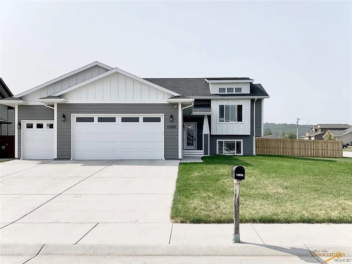 The exterior of a modern two-story home is displayed, featuring a light grey and white color scheme. A spacious driveway leads to an oversized two-car garage with three large garage doors. The well-maintained lawn extends in front, complemented by a fence in the background.