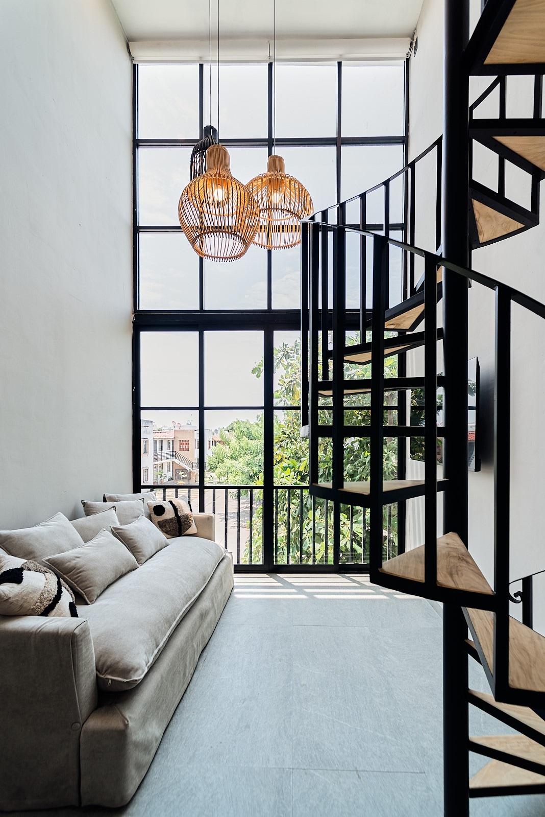 A light-filled living area features large windows, allowing natural light to illuminate the space. A comfortable sofa, adorned with decorative pillows, sits next to a stylish spiral staircase crafted from metal and wood. Pendant lights hang from the ceiling, adding a modern touch.