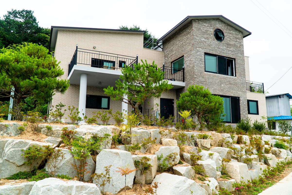 The modern villa features a combination of light-colored bricks and large windows, surrounded by a landscaped garden with various shrubs and rocks. A balcony provides outdoor space on the upper level, and a wide entrance is marked by white columns, leading to a welcoming atmosphere.