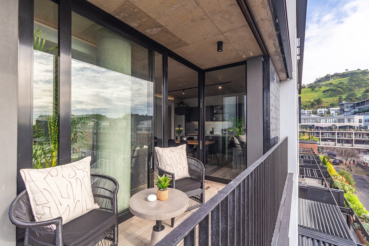 A spacious balcony offers two stylish chairs and a round table, complemented by potted greenery. Large glass doors provide a view into the modern living area, while the surrounding hills and vibrant architecture are visible in the background.
