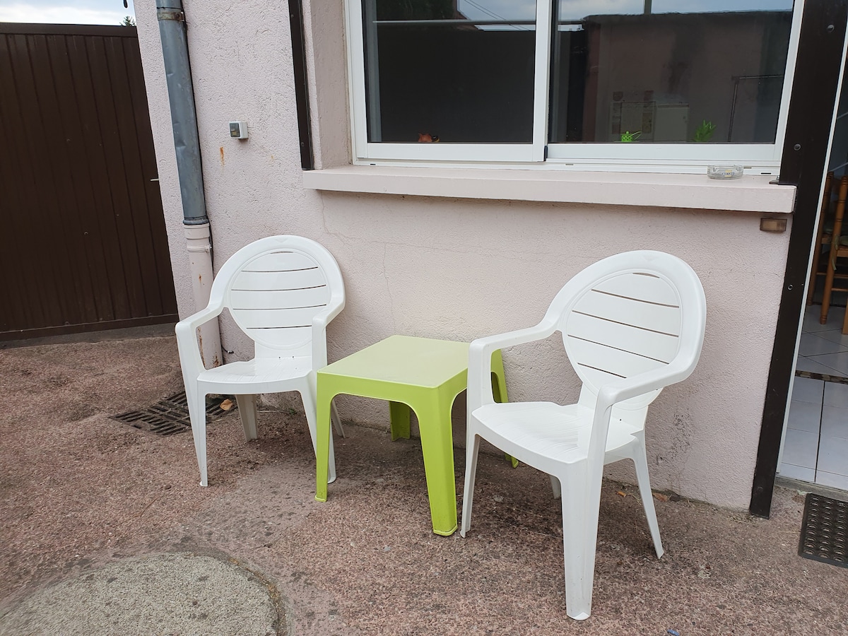 Two white plastic chairs and a small green table are positioned outside against a light-colored wall. The setting provides a simple space for relaxation, with a window visible in the background, allowing for outdoor enjoyment.