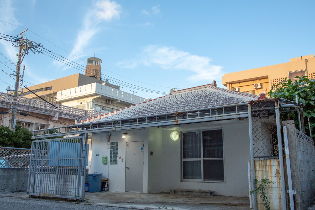 A renovated Okinawan house features a distinct tiled roof and a white exterior. The entrance is framed by a gate, allowing a view of a large window and a security light mounted on the wall. The surrounding area is urban, with nearby buildings visible in the background.