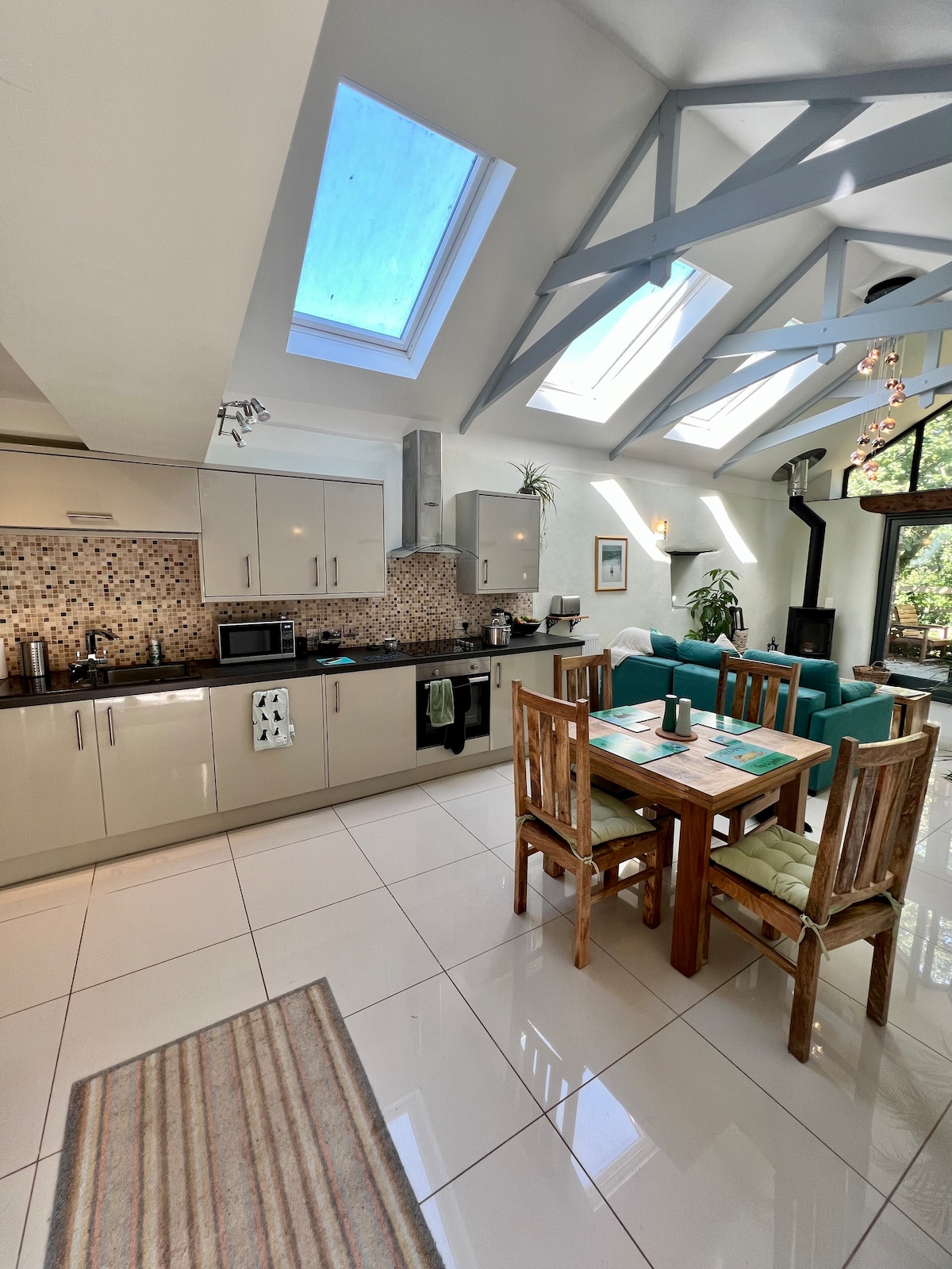A bright and open kitchen features modern cabinetry and appliances, with a dining table set for four. Natural light floods through skylights and windows, highlighting the spacious layout. A seating area with teal cushions complements the room's design, while a log burner provides warmth.