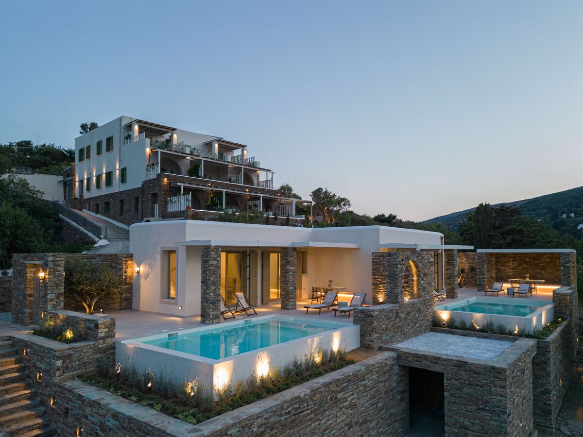 A modern bungalow is set against a twilight backdrop, featuring a private swimming pool surrounded by stone walls. Lounge chairs are positioned alongside the pool. The multi-story structure in the background showcases a sleek design with spacious terraces and greenery integrated into the landscape.
