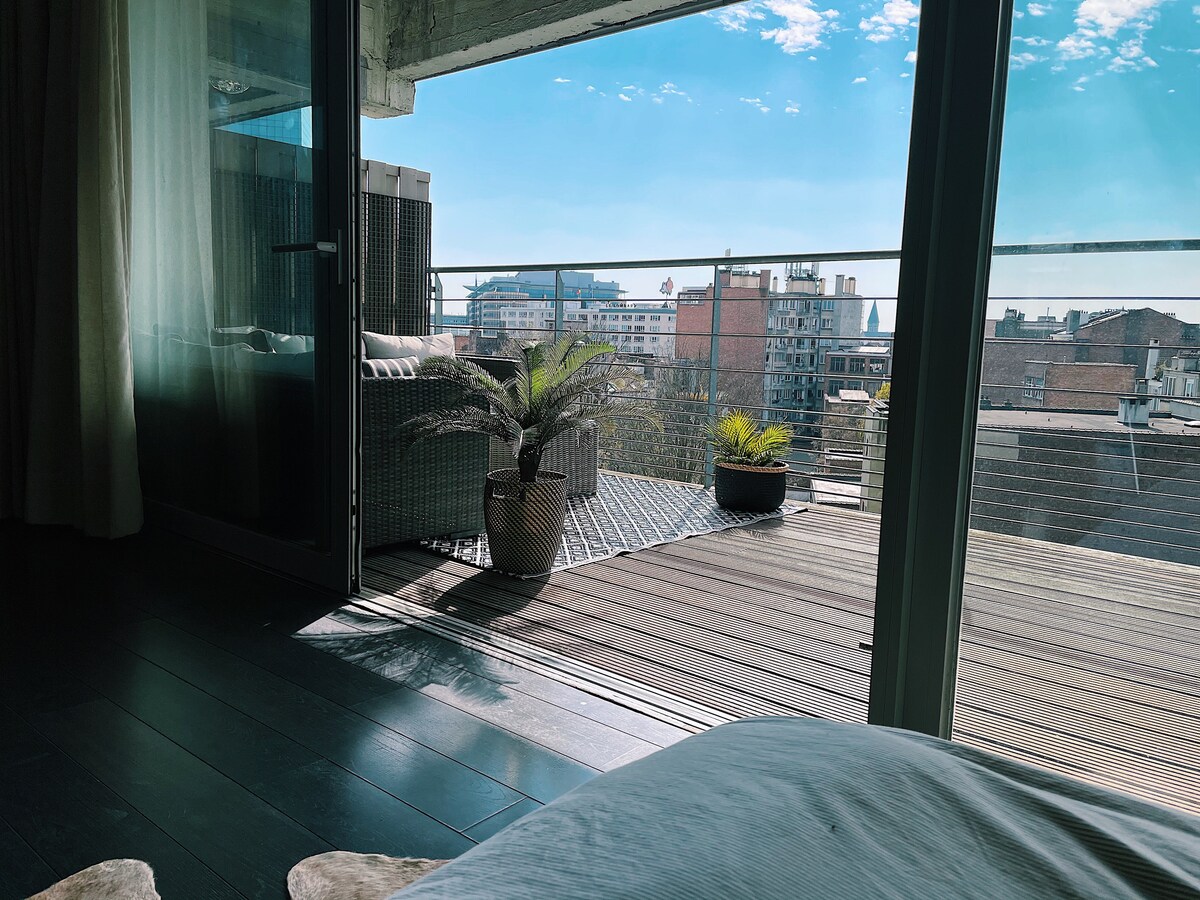 A bright terrace offers a view of the city skyline, featuring potted plants and comfortable seating. Floor-to-ceiling glass doors create a seamless transition from the interior space, allowing natural light to fill the room.