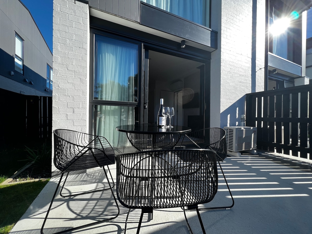 A patio area features a round black table surrounded by four wire chairs. Sunlight creates a warm ambiance over the space, with greenery visible in the background. Large glass doors lead into the townhouse, providing access to indoor living.