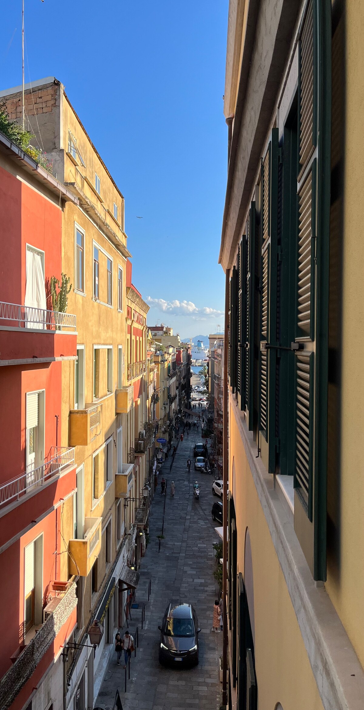 A narrow street is observed from a window, flanked by colorful buildings in warm tones. The view extends to the distant hills and water, with people strolling leisurely along the cobbled path. Shuttered windows add architectural detail to the vibrant scene.