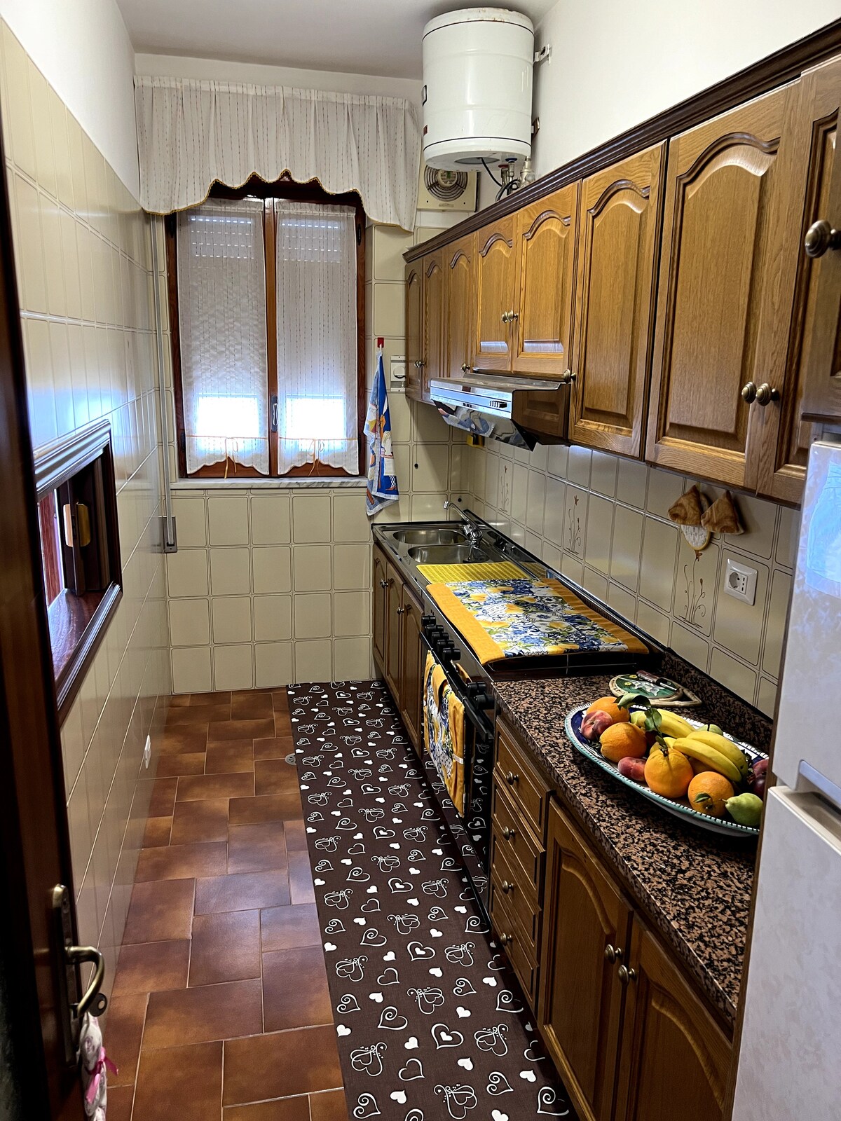 A compact kitchen features wooden cabinets and a tiled backsplash. The countertop is adorned with a fruit bowl filled with a variety of fresh fruits. Natural light enters through a window adorned with a sheer curtain, with patterned tiles covering the floor.
