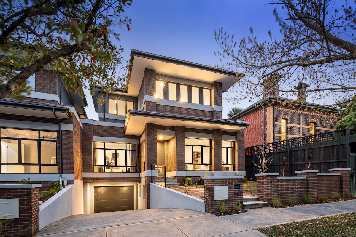 A modern two-story residence is showcased, featuring a spacious front porch supported by brick columns. Large windows create an open and airy feel. The driveway leads to an integrated garage beneath the main structure, surrounded by landscaped greenery and trees.