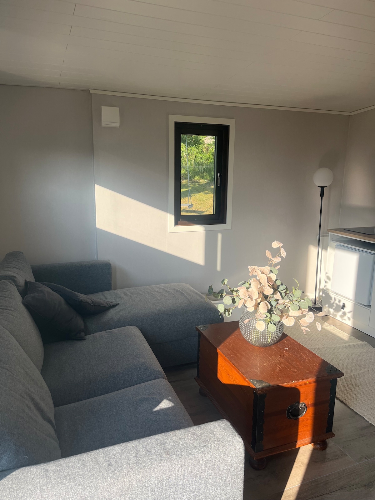 A cozy living area features a spacious gray sofa positioned beside a small wooden table. Natural light filters through a nearby window, illuminating a decorative vase filled with greenery. A floor lamp stands in the corner, providing additional lighting.