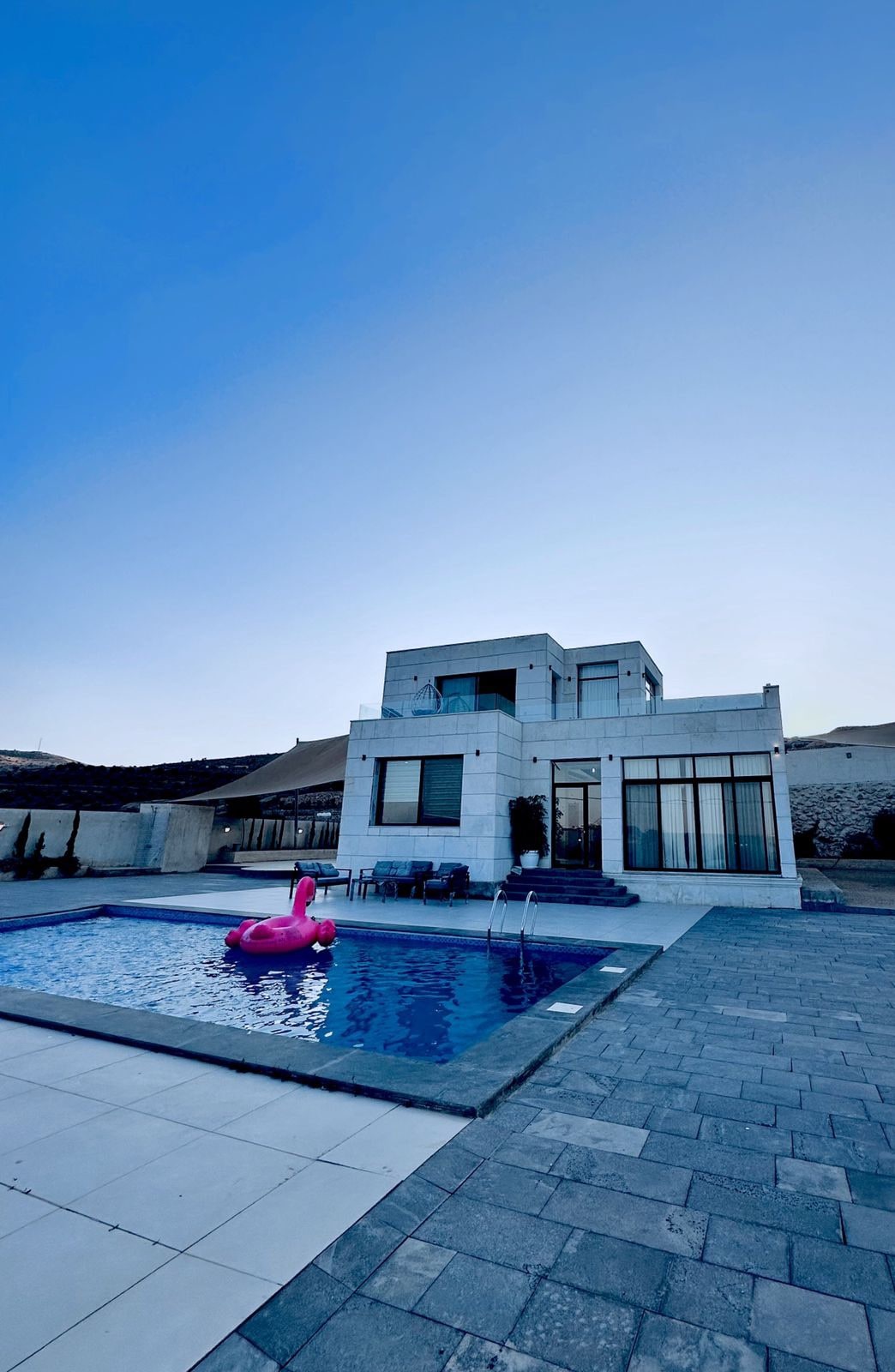 A modern two-story chalet stands beside a serene pool area, featuring a large stone deck. A pink inflatable float rests in the water, while seating is arranged near the pool. The elegant architecture is complemented by clear blue skies in the background.