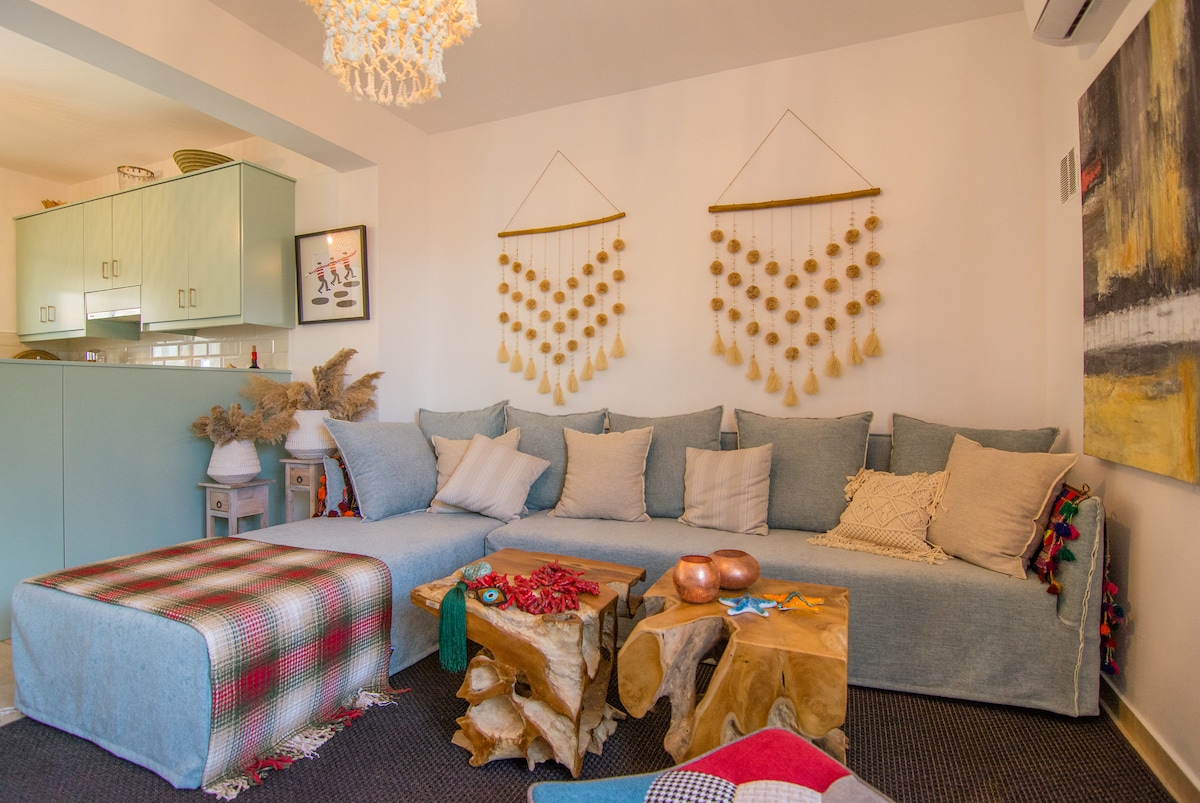 A cozy lounge area features a large, L-shaped sofa adorned with various decorative cushions. Unique wooden tables and a soft blanket add texture, while two artistic wall hangings enhance the eclectic decor. The space is illuminated by a stylish light fixture, creating a welcoming atmosphere.