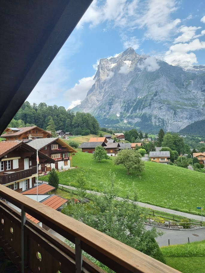 A scenic view from a balcony features lush green meadows and traditional Swiss chalets nestled in the valley. Majestic mountains rise in the background, partially shrouded in clouds, showcasing the natural beauty of the surrounding area.