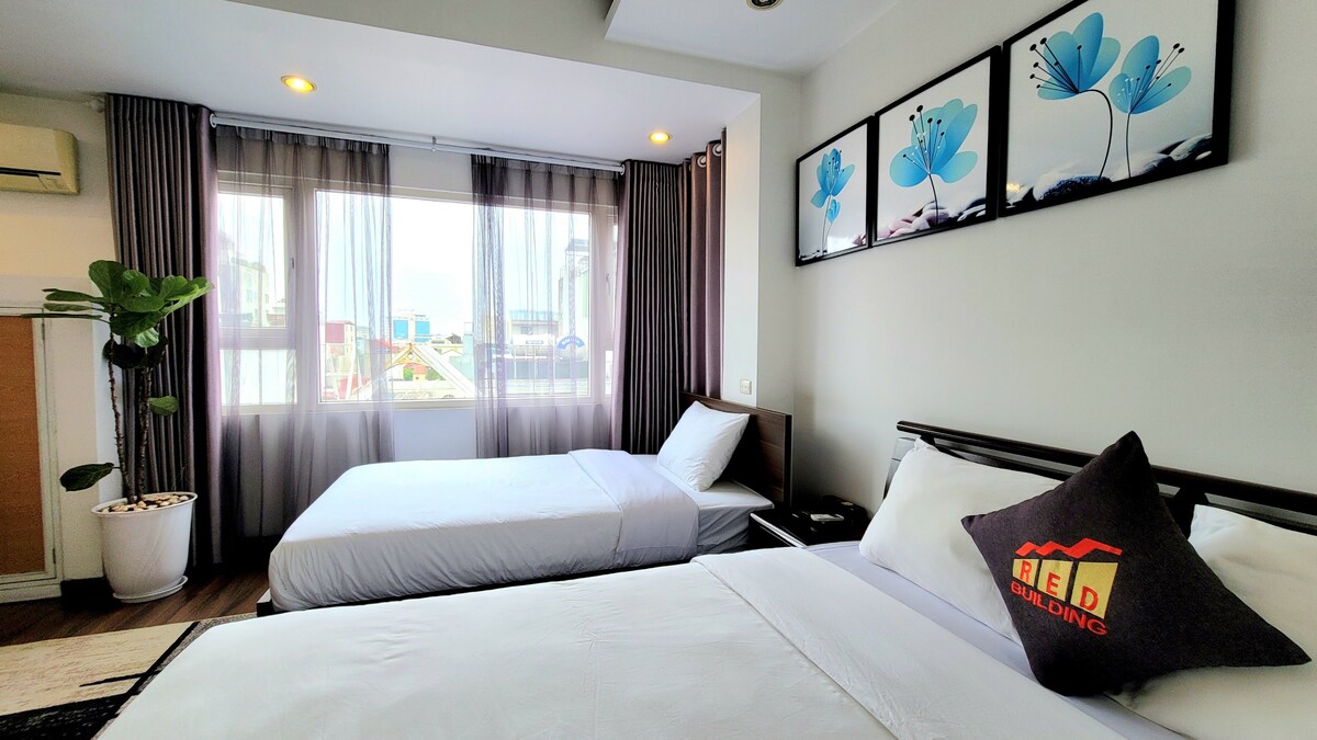 A well-lit room features two beds, providing a spacious sleeping area. Floor-to-ceiling windows allow natural light to fill the space, while sheer curtains add a soft touch. Decorative wall art with floral designs complements the modern aesthetic.
