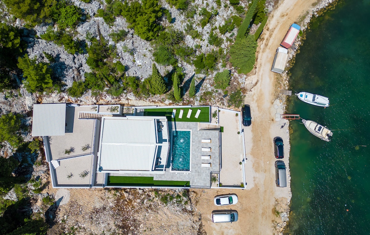 An aerial view of a contemporary four-bedroom house is displayed, featuring a large bio-pool surrounded by lounge chairs. The property is situated near a rocky shoreline with boats docked along the water's edge, providing clear access to the beach.