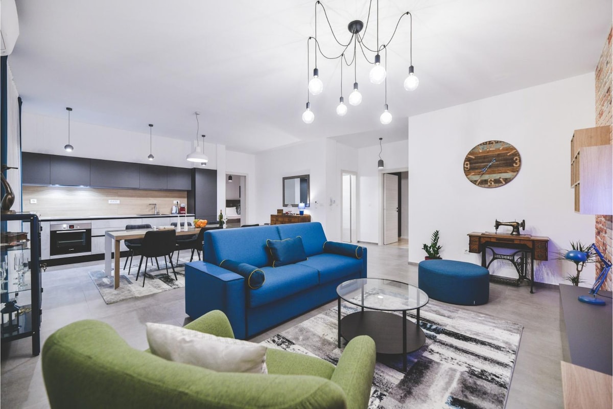 A modern living space features a blue sofa and matching ottoman, complemented by a glass coffee table. The area is illuminated by a hanging light fixture, while a wall clock adds a touch of elegance. A kitchen area with sleek cabinetry is visible in the background.