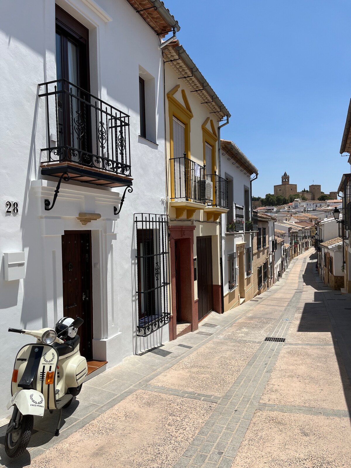 A narrow street lined with traditional white houses featuring decorative balconies and wooden doors. A vintage scooter is parked alongside the building. In the background, a glimpse of a church tower is visible, complementing the charming ambiance of the neighborhood.