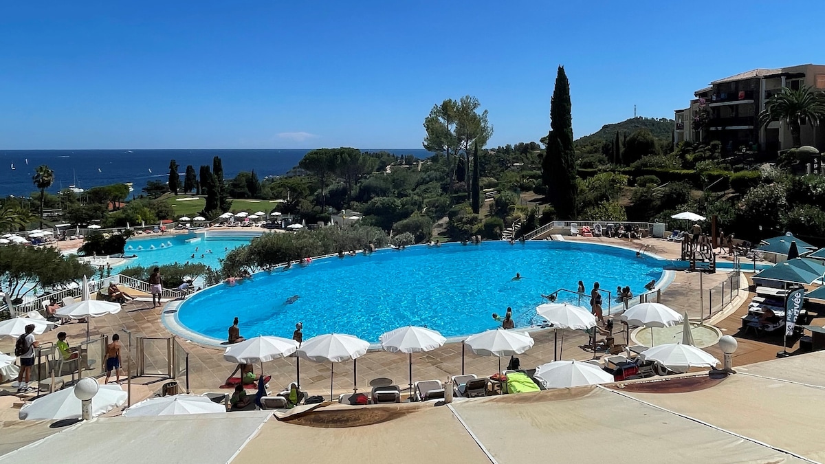 A well-maintained outdoor pool area is visible, featuring a large blue pool surrounded by sun loungers and white umbrellas. Lush greenery and palm trees provide a backdrop, while the sea is seen in the distance under a clear blue sky.