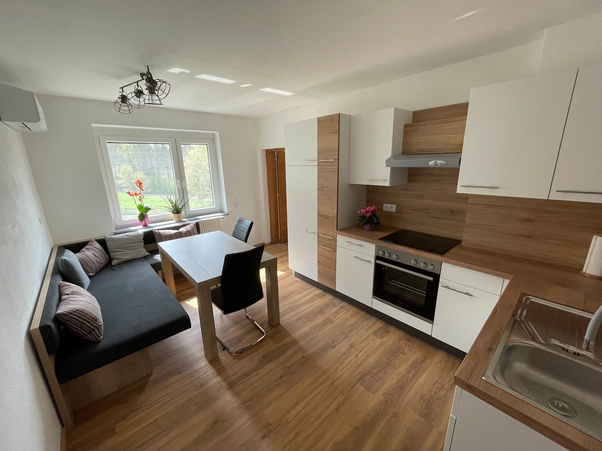 A practical kitchen area features modern cabinetry, a dining table with seating for four, and a cozy seating nook. Natural light filters through a large window, highlighting the stainless steel appliances and wood accents throughout the space.