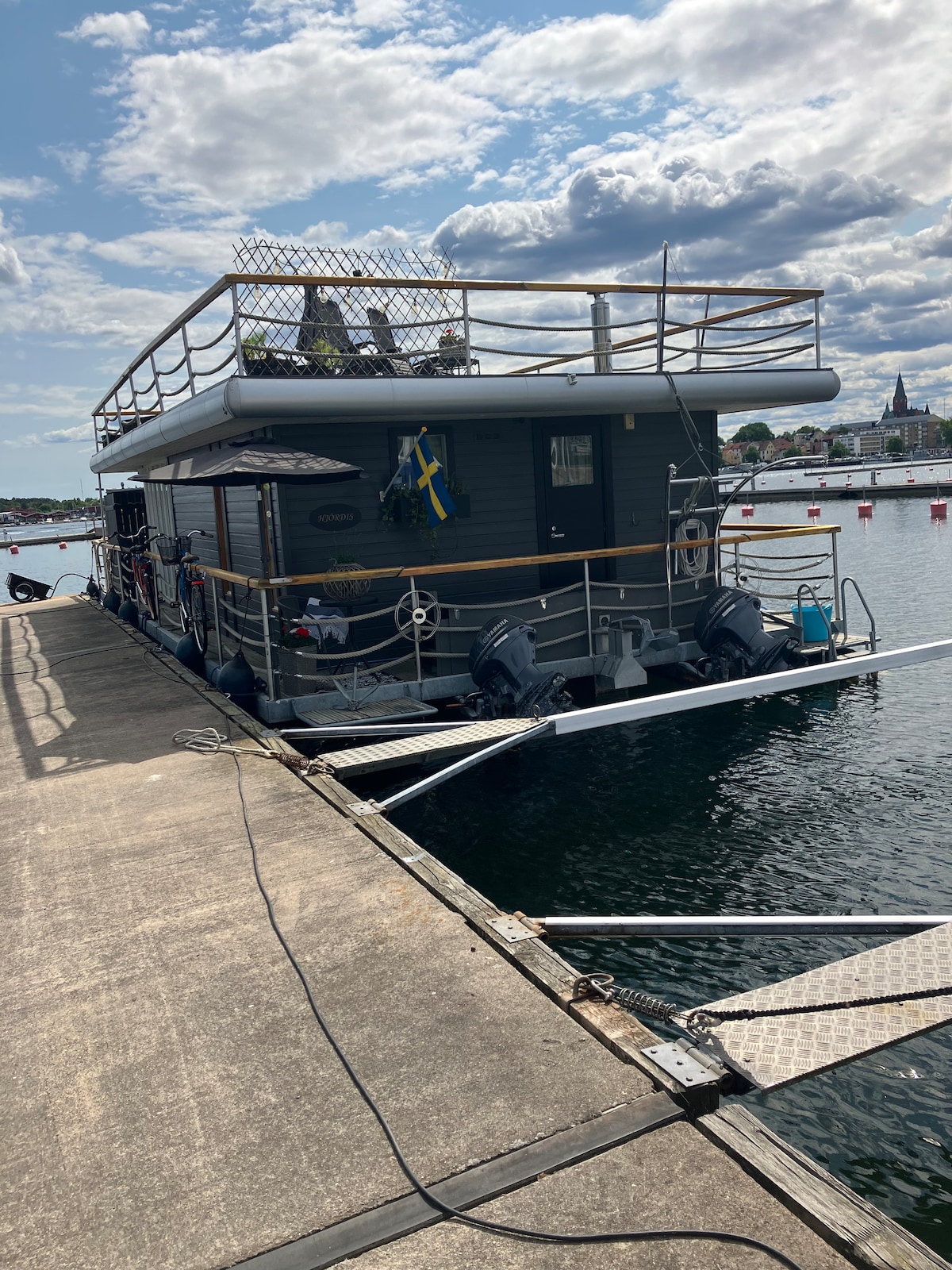 A houseboat is moored alongside a wooden dock, with a spacious upper deck surrounded by railing. The tranquil water reflects the sky and nearby boats. Details such as various decorations and outdoor seating can be seen on the deck, enhancing the unique waterfront experience.