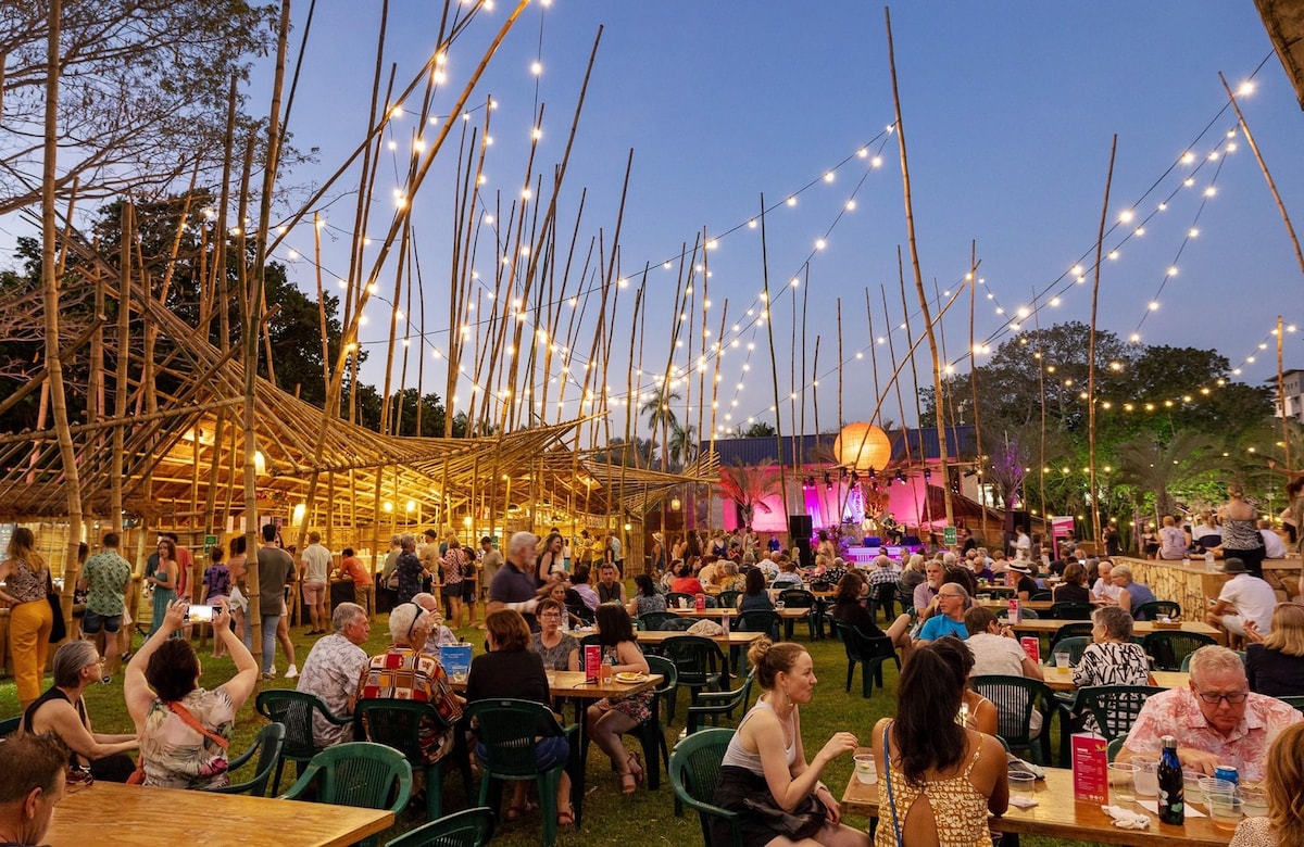 An outdoor gathering space is filled with visitors seated at tables, enjoying an evening event. The area is adorned with tall bamboo structures and string lights, creating a festive ambiance. A stage is visible in the background, showcasing a performance under the dusk sky.