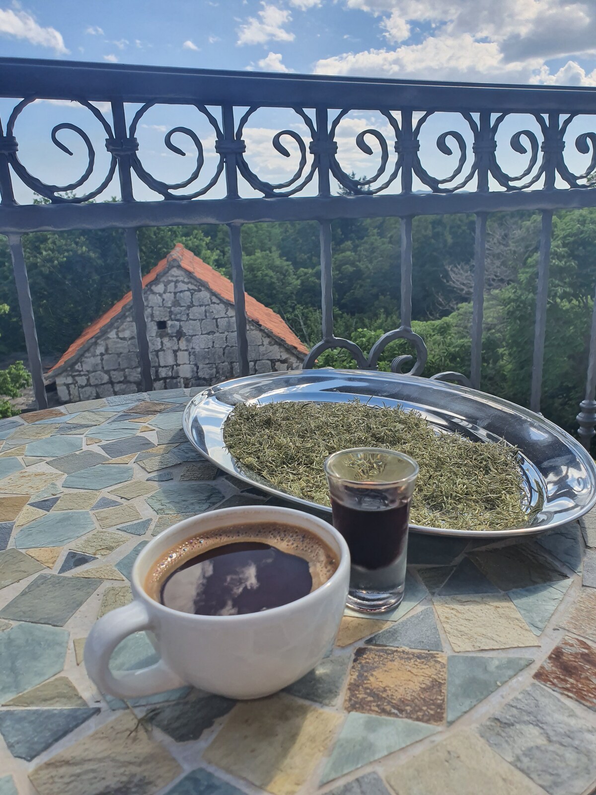A round table covered with a mosaic pattern displays a white cup of coffee beside a small glass of dark beverage. A silver tray rests on the table, holding a collection of dried herbs. A view of green trees and a stone building is seen in the background.