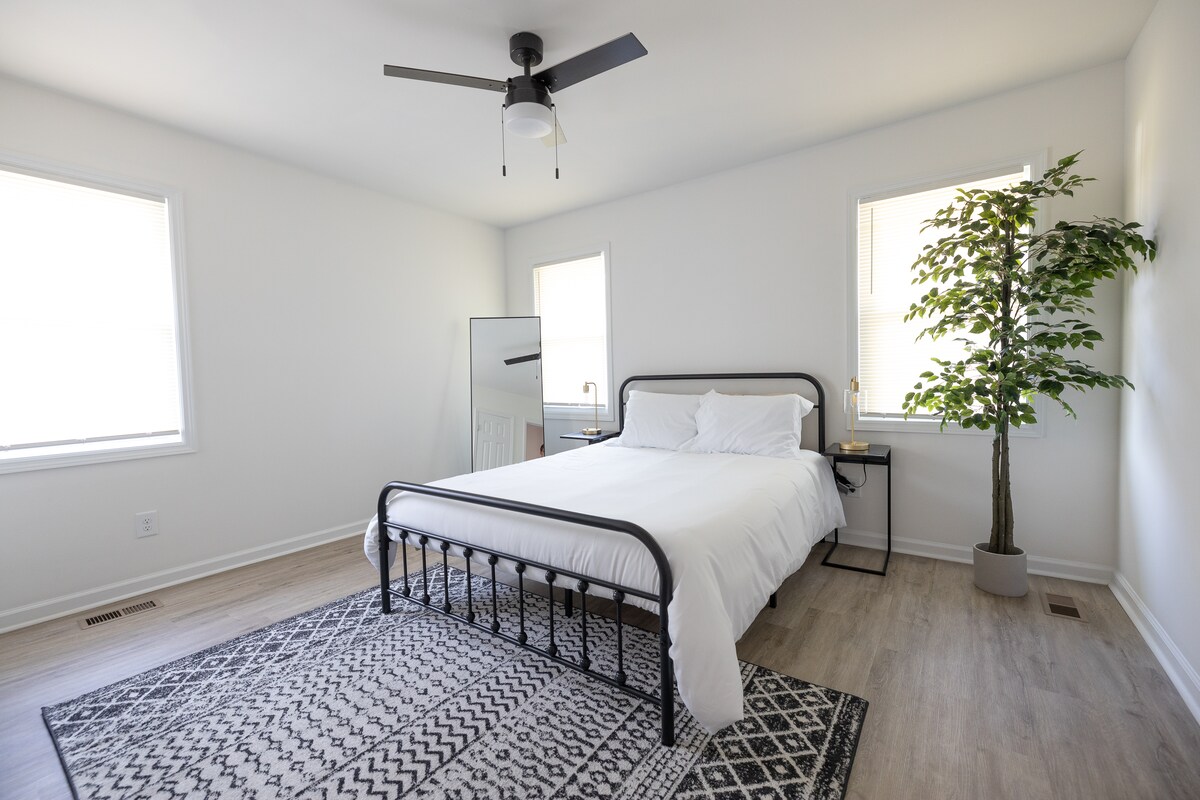 A bright bedroom features a spacious bed with crisp white linens, framed by two windows allowing natural light. A mirror stands against the wall, reflecting the room's simplicity. A potted plant adds a touch of greenery, complementing the neutral color scheme.