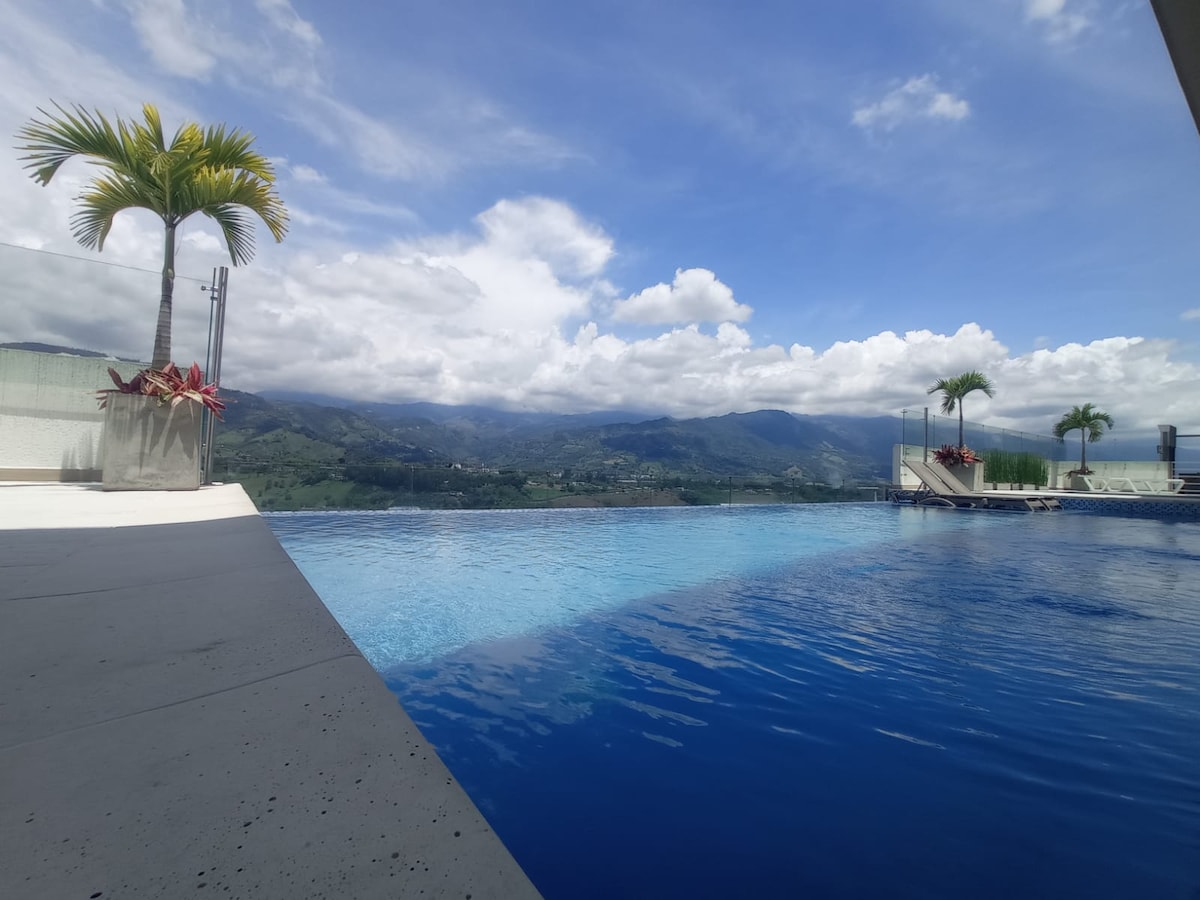 An inviting infinity pool stretches towards the horizon, framed by lush palm trees. Mountains in the distance create a serene backdrop, while fluffy clouds float in a vibrant blue sky. The pool's surface reflects the peaceful surroundings, enhancing the tranquil atmosphere.
