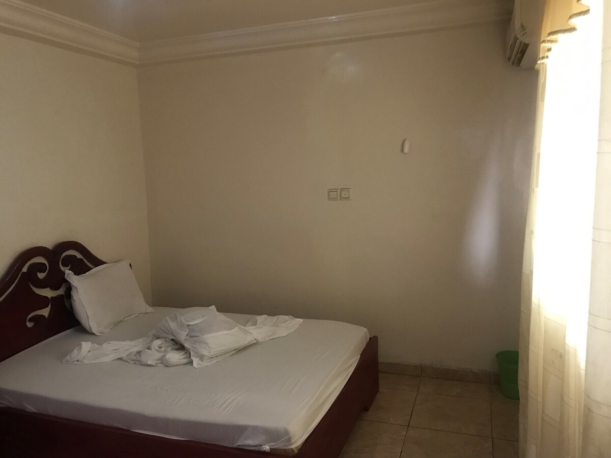 A simple sleeping area features a spacious bed with a white mattress and a single white pillow. Sunlight filters through a sheer curtain, illuminating the room's neutral walls and tiled floor. A small green bin is visible in the corner, maintaining a clean environment.
