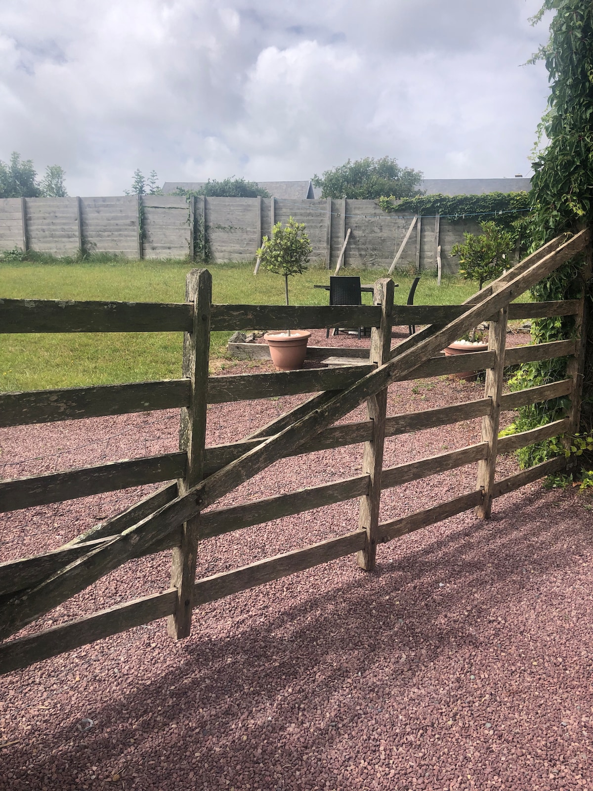 A rustic wooden gate opens to a well-maintained garden area featuring a gravel path. In the background, a small seating arrangement is visible beside potted plants and a young tree. The space is enclosed by a low wall and a lush green landscape.