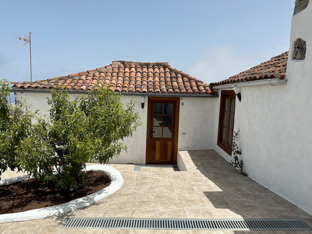 A traditional Canarian house is showcased with a terracotta tiled roof and a welcoming wooden door. Surrounding greenery and a neatly paved pathway lead to the entrance. Windows complement the structure, allowing natural light to illuminate the exterior.