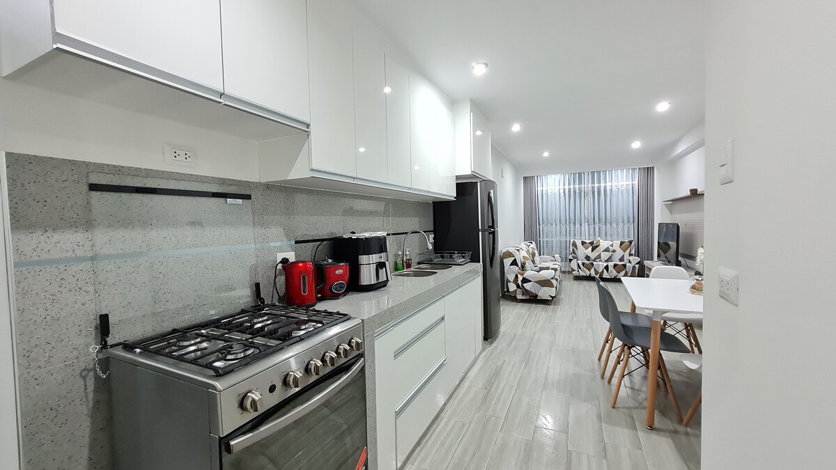 A modern kitchen is visible, featuring sleek cabinets and a spacious countertop. Stainless steel appliances include a gas stove and a refrigerator. A dining area is present, equipped with a white table and several chairs. Natural light flows in through large windows adorned with light curtains.