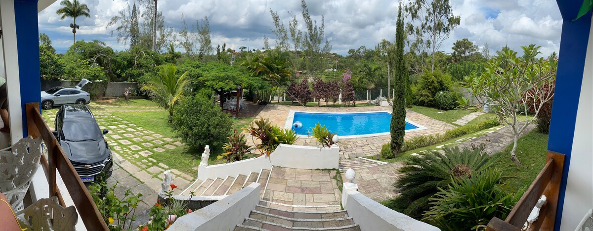A panoramic view of the outdoor space captures a swimming pool surrounded by lush greenery and a well-maintained garden. Stone pathways lead from the steps of a home to the pool area, with various trees and plants framing the scene under a partly cloudy sky.